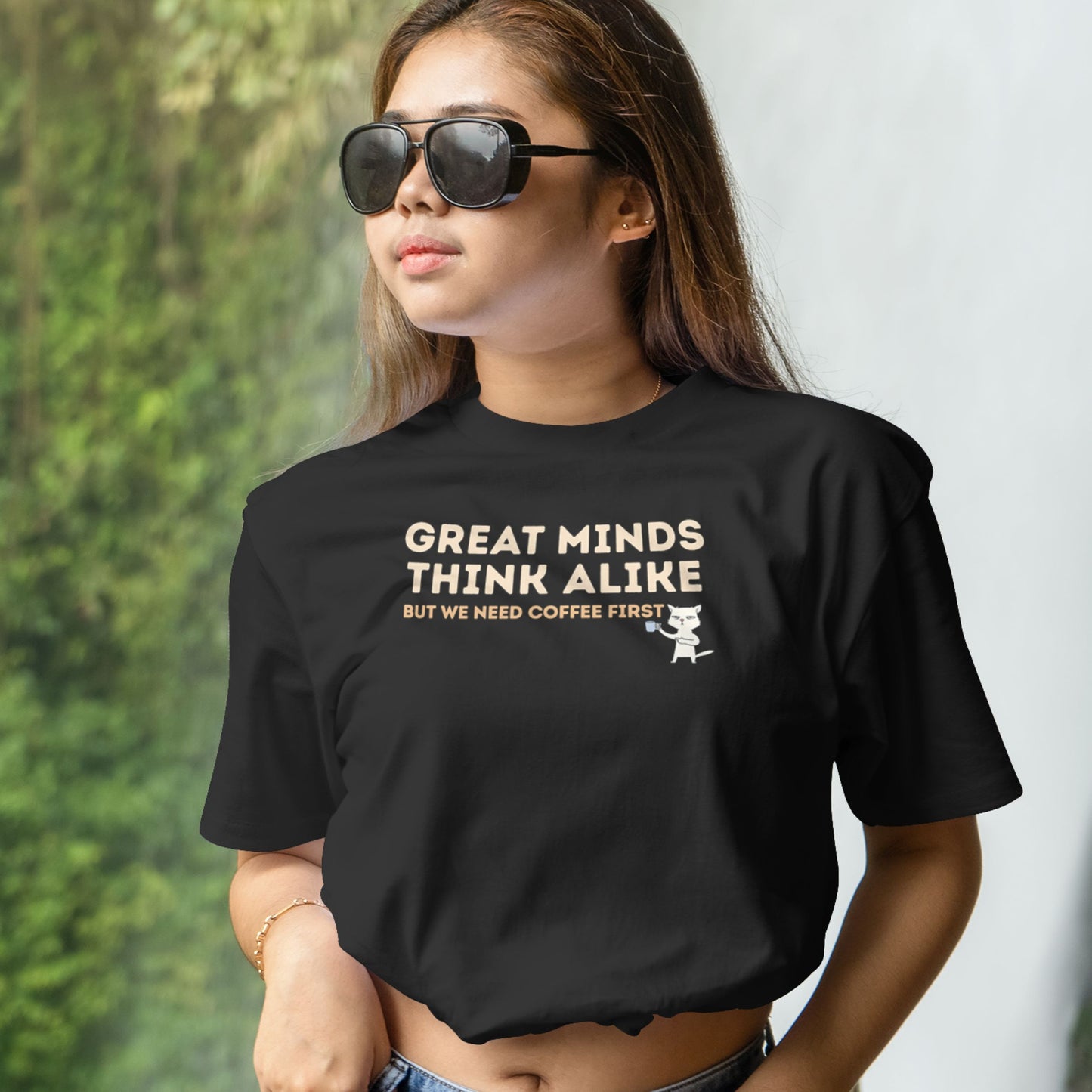 Angled view of a black graphic T-shirt with the quote "Great Minds Think Alike, But We Need Coffee First" and a coffee-loving cat illustration.