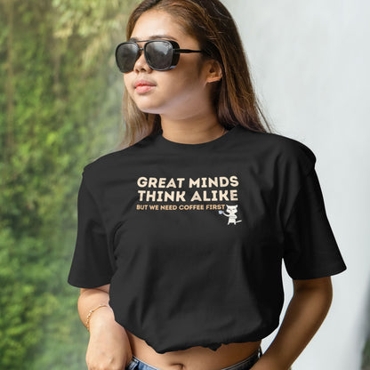 Angled view of a black graphic T-shirt with the quote "Great Minds Think Alike, But We Need Coffee First" and a coffee-loving cat illustration.