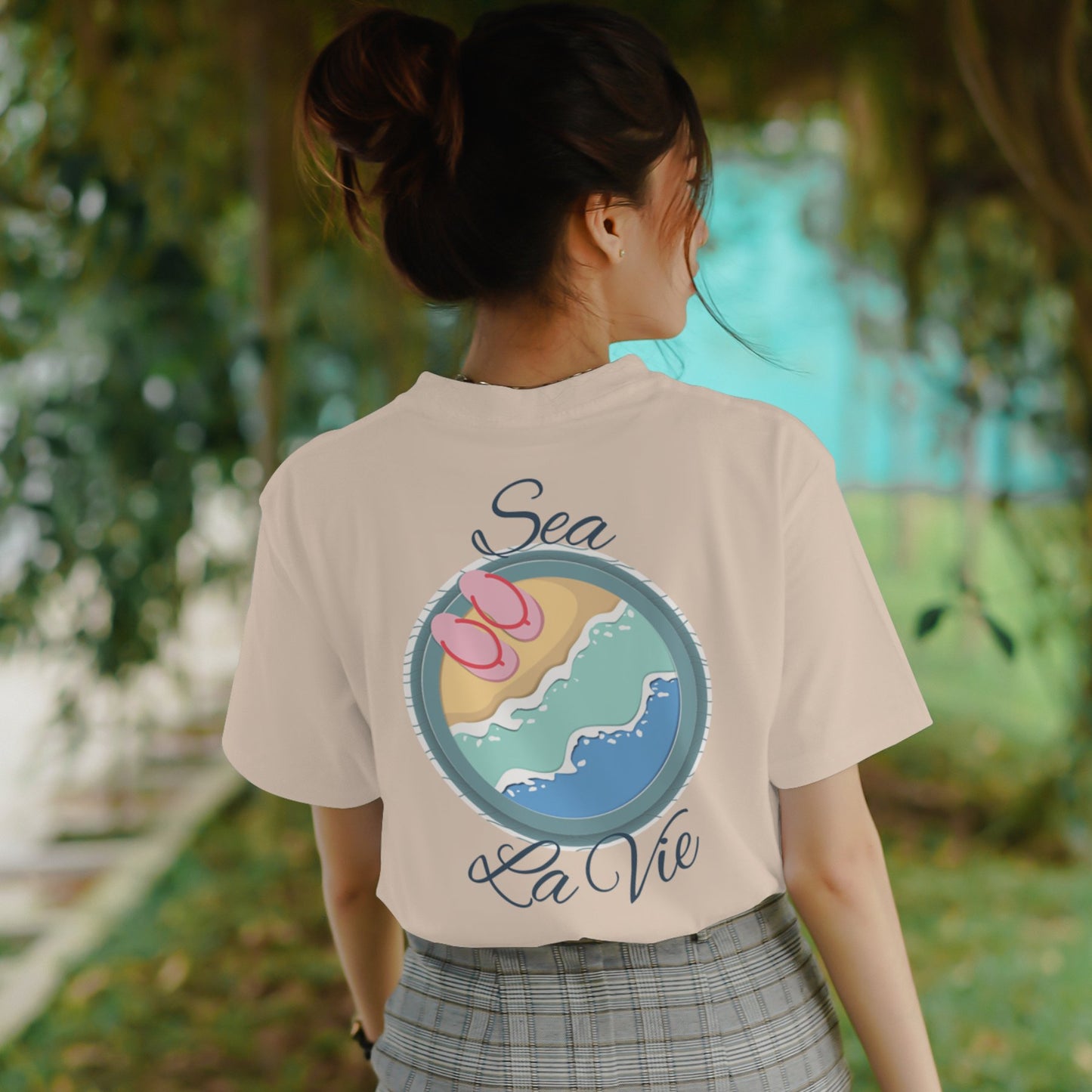 Woman wearing beige oversized travel t-shirt with beach print and quote “Sea La Vie.”